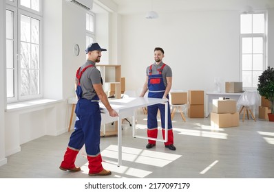 Loaders Carrying Furniture In Flat. Two Male Moving Service Workers Move Furniture When Moving To Office Or Apartment. Young Active Male Loaders In Blue And Red Overalls Carry White Desk.
