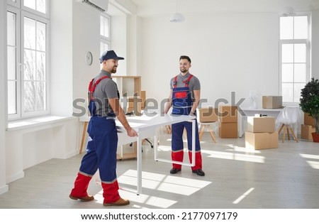 Loaders carrying furniture in flat. Two male moving service workers move furniture when moving to office or apartment. Young active male loaders in blue and red overalls carry white desk.