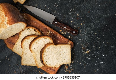 Loaf Of Fresh Sliced White Bread Laid Out On A Cutting Board And A Sharp Knife Nearby. Top View With Copy Space, Bread Crumbs On Black Table Top