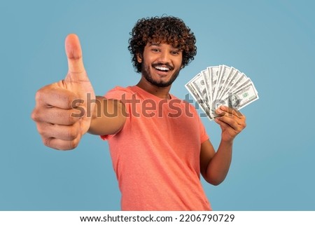 Loan, credit, gambling, trading, bet, lottery concept. Cheerful handsome young indian man in pink t-shirt showing bunch of cash dollars and thumb up over blue studio background