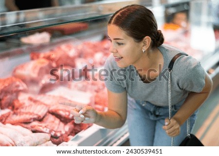 In local butcher shop, customer girl looks thoughtfully at glass display case of refrigerator and chooses pork wing ribs