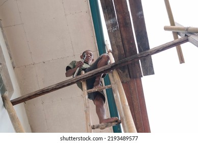 A local fearless carpenter repairing the ceiling of a two story house while sitting on flimsy scaffolding and a ladder and not wearing safety gear.