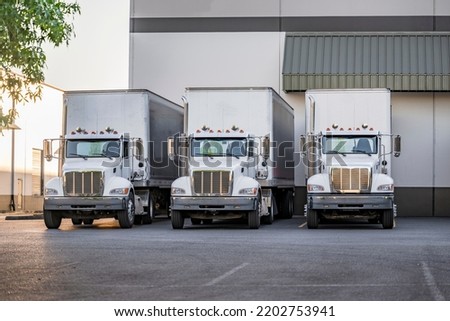 Local haul middle duty professional white big rigs semi trucks tractors with box trailers waiting for the next load standing in row on the warehouse industrial parking lot with green trees