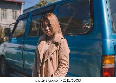 A Local Tourist With Blond Hair And Wearing A Beige Trench Coat Enjoying The Cold Breeze Outdoors With A Blue Van As Background.