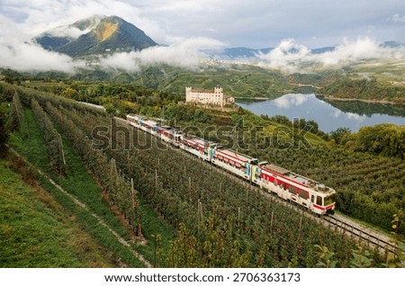 A local train dashing by an apple orchard, beautiful Cles Castle perching by Lake Santa Giustina and majestic mountains towering in background, in Val di Non, Trentino Alto Adige, South Tyrol, Italy