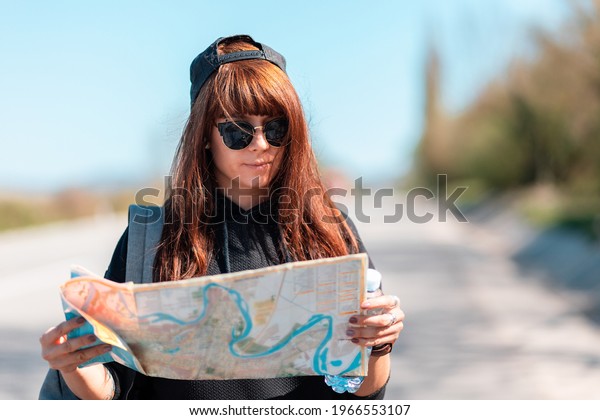 Local Travel. Portrait of young\
woman wearing a cap and sunglasses, holding a paper map. In the\
background, the road is blurred. The concept of\
hitchhiking.