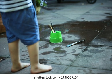 Lockdown Activity For Kids - Mess Free Painting With Water. Unidentified Child Wearing Shorts Stands Near Paint Brushes And Green Containers Painting The Ground With Paint Brushes And Water.