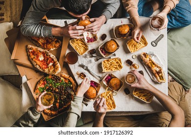 Lockdown Fast Food Dinner From Delivery Service. Flat-lay Of Friends Sitting And Having Beer Quarantine Party With Burgers, French Fries, Sandwiches, Pizza And Salad Over Table Background, Top View