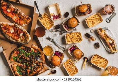 Lockdown fast food dinner from delivery service concept. Flat-lay of quarantine home party with burgers, french fries, sandwiches, pizza, beer in glasses over white tablecloth background, top view