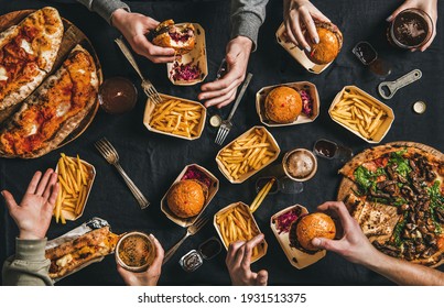 Lockdown fast food dinner from delivery service. Flat-lay of peoples hands eating burger, fries, sandwiche, pizza, drinking beer over table background, top view. Quarantine home party, takeaway food - Shutterstock ID 1931513375