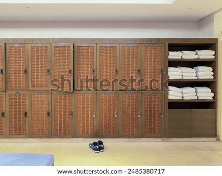 Locker room interior, featuring a row of wooden lockers with wicker fronts. Each locker is labeled with a number, and there are clean, neatly folded white towels stacked on open shelves to the right.