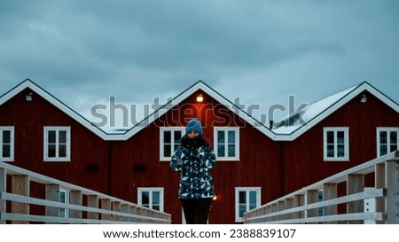 Lofoten islands, Senja region, Norway. Winter. Famous fishing town. Young girl in blue jacket and hat stands on bridge, behind red fishing houses. Concept of tourism.