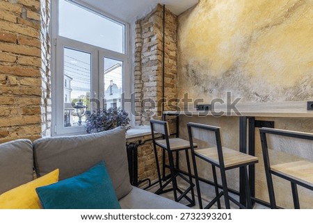 Loft hostel common area in daylight. Features wooden bar stools at a high counter against a textured yellow wall. A large window with brick trim reveals a classic street view, providing a bright and inspiring workspace for guests.