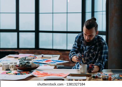 Loft studio working space. Creative process. Young talented artist painting his abstract artwork, washing brush in white mug.