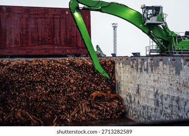 Log loading process at a seaport terminal. A green port material handler equipped with a grab loads roundwood logs into the cargo hold of a vessel. Industrial port operations, timber export