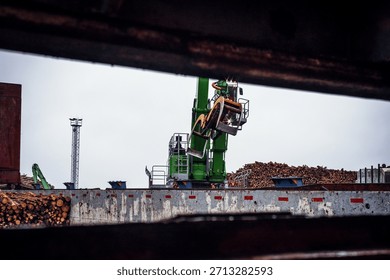 Log loading process at a seaport terminal. A green port material handler equipped with a grab loads roundwood logs into the cargo hold of a vessel. Industrial port operations, timber export