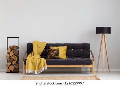 Log of wood next to comfortable sofa with yellow blanket and pillows, stylish wooden lamp with black lampshade, real photo copy space on the empty grey wall