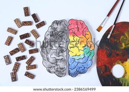 Logic and creativity. Paper brain with one colorful part and another grey on white background, flat lay. Domino tiles near left hemisphere and palette with brushes near right one