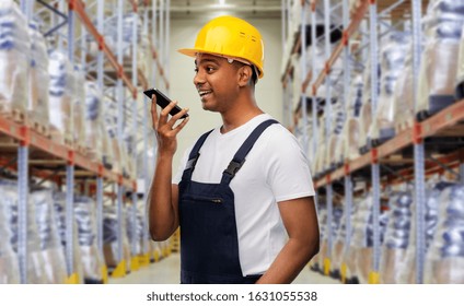 Logistic Business, Technology And People Concept - Happy Smiling Indian Loader Or Worker In Helmet Using Voice Command Recorder On Smartphone Over Warehouse Background