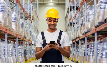 logistic business, technology and people concept - happy smiling indian loader or worker in helmet with smartphone over warehouse background