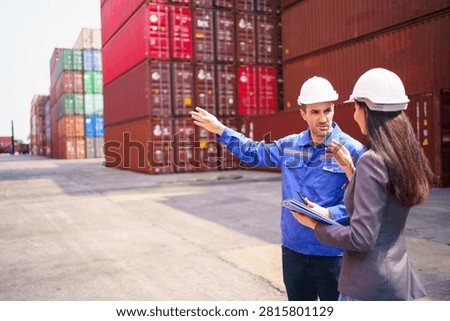 Logistics engineer in blue uniform and white hardhat pointing hand to explain container layout to businesswoman while holding clipboard at shipping cargo container yard during inspection walkthrough.