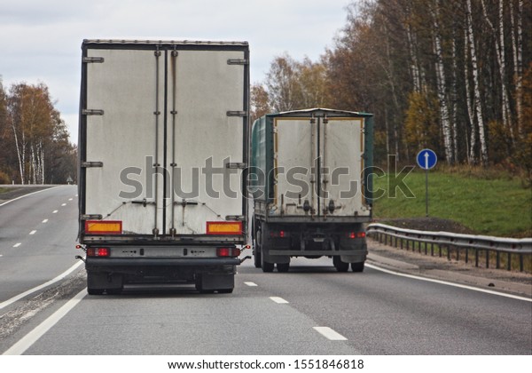 Logistics industry,\
international freight by road, two white trucks drive on all\
two-lane asphalted country road in autumn day, rear view,\
overtaking rules for\
drivers