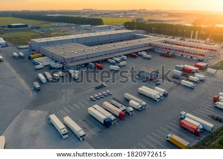 Logistics park with warehouse, loading hub and many semi trucks with cargo trailers standing at the ramps for load/unload goods at sunset. 