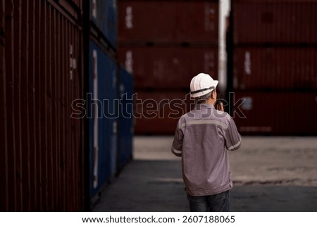 A logistics supervisor in a hard hat and uniform communicates via radio while holding a clipboard at a busy shipping yard. Industrial operations and transportation management in action.