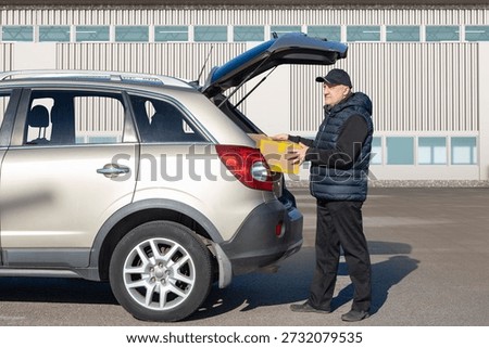 Logistics worker loads a cardboard box into an SUV trunk. Package handling and vehicle loading by a delivery courier. Transportation and distribution services.