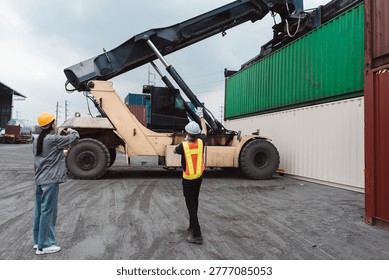 Logistics workers directing reach stacker lifting cargo container. Industrial engineer and operator managing heavy equipment transport at shipping yard.