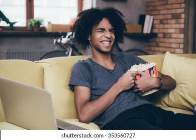 LOL. Cheerful Young African Man Watching TV And Holding Bucket Of Popcorn While Sitting On The Couch At Home