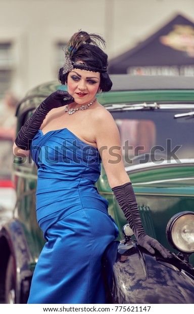 london, england,\
02/05/217, A gatsby girl in 1920s Art deco style party girl flapper\
clothing, and feather head wear, black gloves and blue dress, \
posing by a  vintage retro car.\

