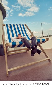 London, England, 05/05/2017, A Stylish Retro Vintage Fashionable Man Sat Relaxing With Arms On Head, On A Giant Retro Seaside Deckchair. Giant Deckchair. Best Dressed Award. 