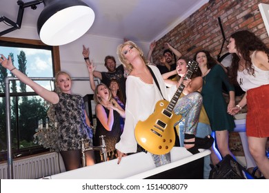 London, England 05/05/2018 Fancy Dress Party, Woman In A Glam Rock Sequin Costume In A Bathtub With A Guitar Posing. Pretending To Be Playing In A Music Concert At A House Party.