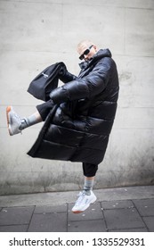 LONDON - FEBRUARY 17, 2019: London Fashion Week. Blonde Man Wears Clear Pvc Belt Bag, Black Down Jacket And Grey Trousers, White Sneakers With Saunt Lauren Bag