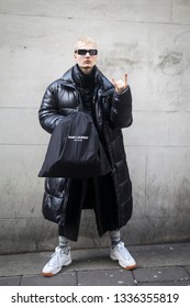 LONDON - FEBRUARY 17, 2019: London Fashion Week. Blonde Man Wears Clear Pvc Belt Bag, Black Down Jacket And Grey Trousers, White Sneakers With Saunt Lauren Bag