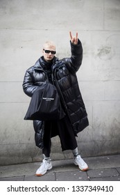 LONDON - FEBRUARY 17, 2019: London Fashion Week. Blonde Man Wears Clear Pvc Belt Bag, Black Down Jacket And Grey Trousers, White Sneakers With Saunt Lauren Bag