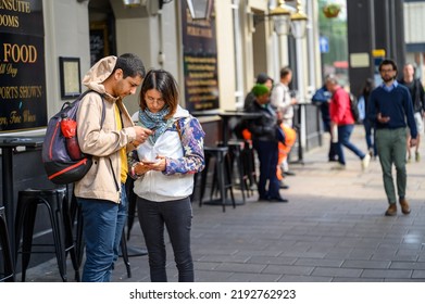 LONDON - May 20, 2022: Man And Woman With Backpacks Looking At Smartphones On Busy London Street