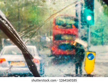 London Street Scene Abstracted By Rain On A Car Windscreen On A Rainy Day. A Red Bus Can Be Seen And A Man Holding Cardboard Above His Head For Shelter By A Yellow Arrow Indicating A Road Island.
