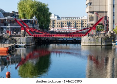 London. UK- 05.30.2020: A Red Painted Pedestrian Draw Bridge In St. Katherine Docks Marina Casting Reflection On The Water With Building On The South Bank Of The Thames River In The Background.