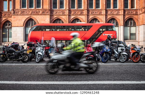 LONDON, UK - 13 SEPTEMBER 2018: A roadside\
motorcycle parking lot in the heart of the City of London with a\
motorcycle passing the frame and a location specific red London bus\
in the background.