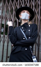 London, UK - 15 February, 2020 People wear mask for protection during London fashion week. Street style. Stylish woman in black dress, coat and a wide-brimmed hat wears a beige mask. Fashion week