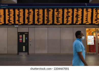 London, UK - 16 July, 2022 - Digital Train Timetable At London Bridge Station, With A Masked Passenger Walking Past