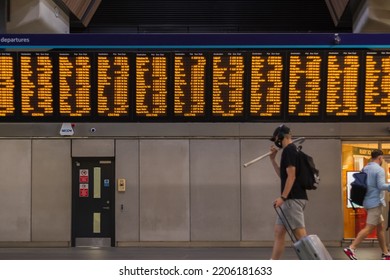 London, UK - 16 July, 2022 - Digital Train Timetable At London Bridge Station, With Masked Passenger Walking Past