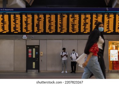 London, UK - 16 July, 2022 - Digital Train Timetable At London Bridge Station, With Masked Passengers Walking Past