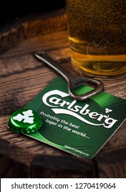 LONDON, UK - AUGUST 10, 2018: Carlsberg beer coaster with bottle top and opener and glass of beer on top of wood barrel.