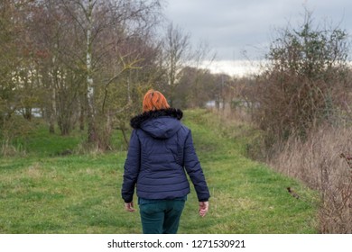 London, UK - Circa January 2019: Middle Aged Woman With Red Hair Seen Walking Away From The Camera, Along A Nature Trail In A Popular Nature Reserve. She Is Seen Wearing A Winter Jacket And Attire.