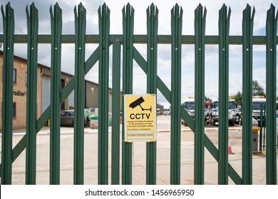 London, UK - Circa July 2019; Shallow focus of a CCTV security sign seen on a metal, secure industrial fence. Beyond the fence is a large warehouse and office buildings, prone to break-ins.