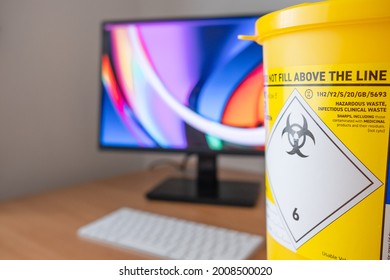 London, UK - Circa July 2021: Close-up, Shallow Focus Of A Medical Sharps Waste Yellow Bin Seen On A Doctors Desk. The Biohazard Symbol Can Be Seen Used For Disposal Of Syringes, Blood Products Etc.