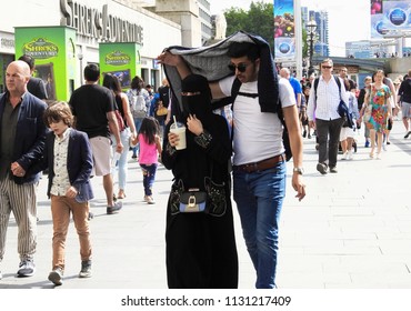 London, UK. July 10 2018. A Muslim Man Protects His Wife - Wearing A Full Black Burka - From The Hot Sun With His Jacket As They Walk Together Along London's South Bank Near Westminster Bridge.